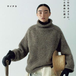 May include: A person wearing a grey turtleneck sweater, black gloves, and holding a straw hat and a wooden tool. The image includes Japanese text. The book title is "Nomad Knit" and the author's name is "Saichika".