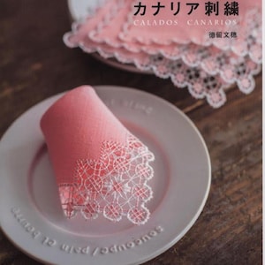 Peut inclure: Un mouchoir rose plié avec une bordure en dentelle blanche, posé sur une assiette gris clair. L'assiette porte du texte sur le bord. Une pile de napperons en dentelle rose similaires est en arrière-plan. Le livre indique "Margarita & Fumiho's Canary Embroidery".
