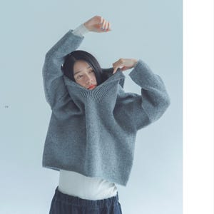 Peut inclure: Un pull gris surdimensionné avec un col côtelé et des manches longues. Le pull est porté sur un t-shirt blanc à manches longues et un pantalon bleu foncé. Les bras du modèle sont levés, et le pull est remonté autour du visage.