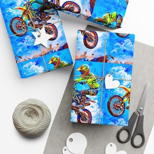 Op de afbeelding: Cadeaupapier met een aquarel illustratie van een motorcrosser op een blauwe achtergrond met wolken. Het cadeaupapier is vastgebonden met touw en versierd met stervormige en hartvormige labels. Een klosje touw en een schaar zijn ook zichtbaar.