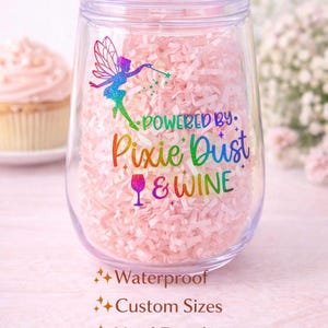 May include: A clear, stemless wine tumbler with a lid and a pink straw. The tumbler is decorated with a colorful fairy design and the words "Powered by Pixie Dust & Wine." The tumbler is filled with pink shredded paper.
