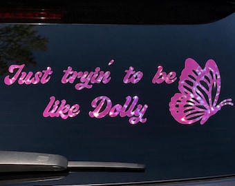 May include: A pink holographic vinyl decal applied to a dark vehicle window. The decal features the text "Just tryin' to be like Dolly" in a cursive script font, accompanied by a detailed butterfly graphic positioned to the right of the text.