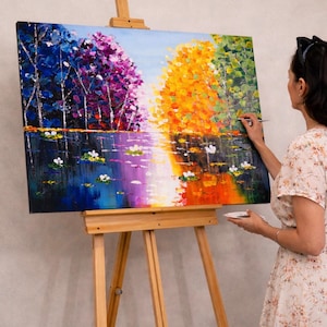 May include: An original oil painting on canvas depicting a colorful landscape with trees and water lilies. The artwork is displayed on a wooden easel. The painting features vibrant hues of blue, purple, orange, and green.