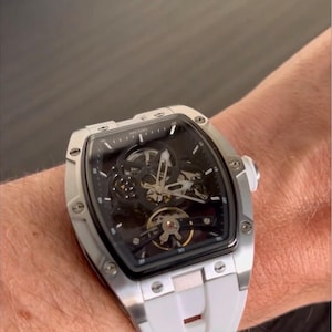 May include: A silver-toned wristwatch with a black face and a white band is shown. The watch has a skeleton design, revealing the inner workings. The watch is on a wrist.
