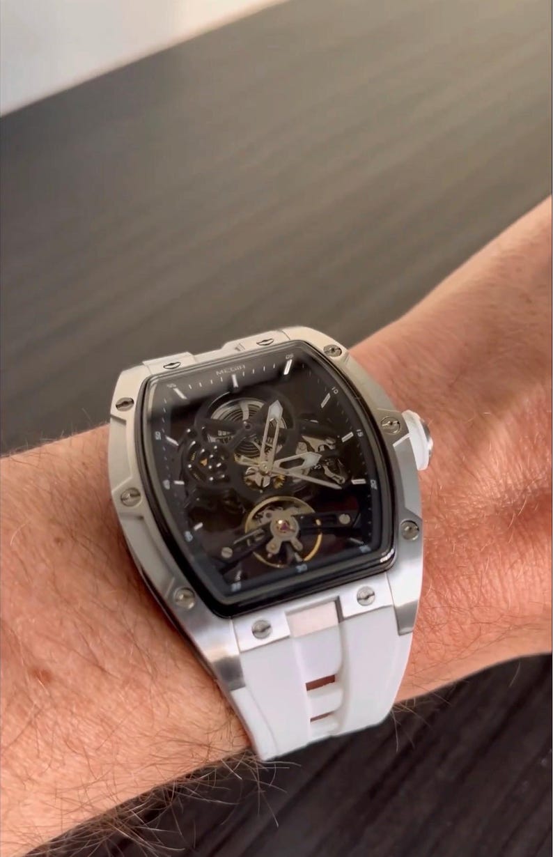 May include: A silver-toned wristwatch with a black face and a white band is shown. The watch has a skeleton design, revealing the inner workings. The watch is on a wrist.