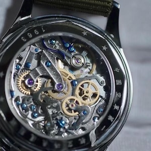 May include: Close-up of a silver wristwatch with a transparent face revealing intricate mechanical gears. The watch features a green fabric strap and the number "1963" is visible on the bezel. The watch's internal components are detailed with blue and gold accents.