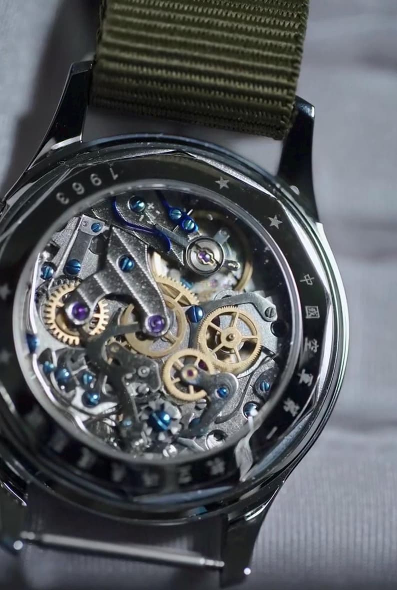 May include: Close-up of a silver wristwatch with a transparent face revealing intricate mechanical gears. The watch features a green fabric strap and the number "1963" is visible on the bezel. The watch's internal components are detailed with blue and gold accents.