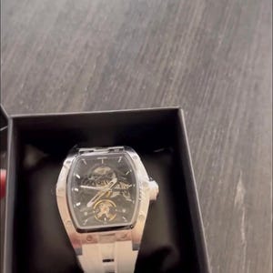 May include: A silver and black wristwatch with a white band is displayed in a black box. The watch has a skeleton dial, revealing the inner workings. The watch has a rectangular face and a white silicone band.