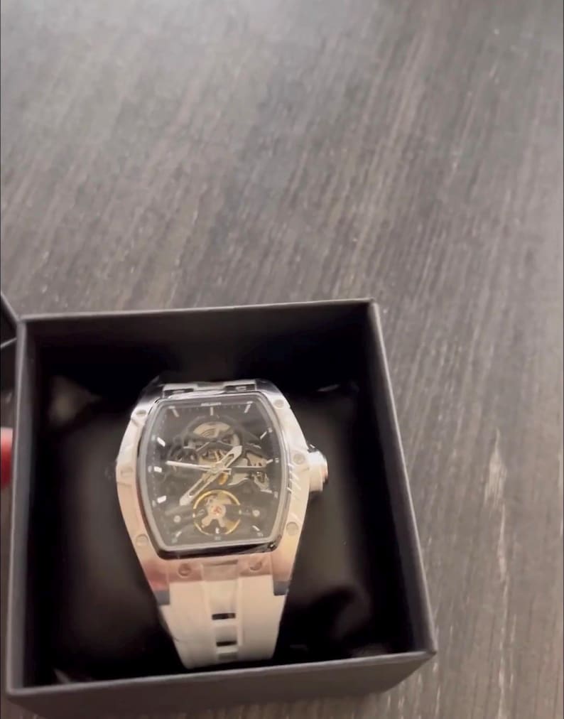 May include: A silver and black wristwatch with a white band is displayed in a black box. The watch has a skeleton dial, revealing the inner workings. The watch has a rectangular face and a white silicone band.