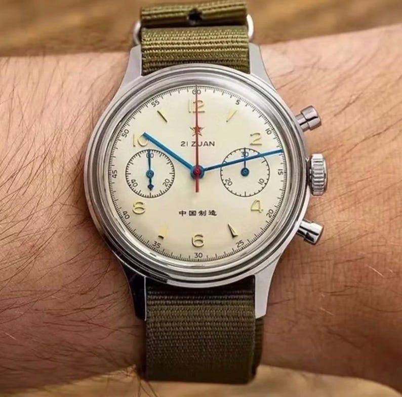 May include: A vintage-style wristwatch with a cream-colored face, featuring blue and red hands, and a green canvas strap. The watch has a silver-colored case and multiple sub-dials. The text "21 ZUAN" and Chinese characters are visible on the face.