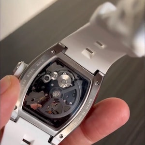 May include: A skeleton watch with a white silicone strap. The watch features a transparent face, revealing the intricate inner workings of the mechanism. The watch case is silver-toned, and the back is marked "Stainless Steel Back" and "Water Resistant".