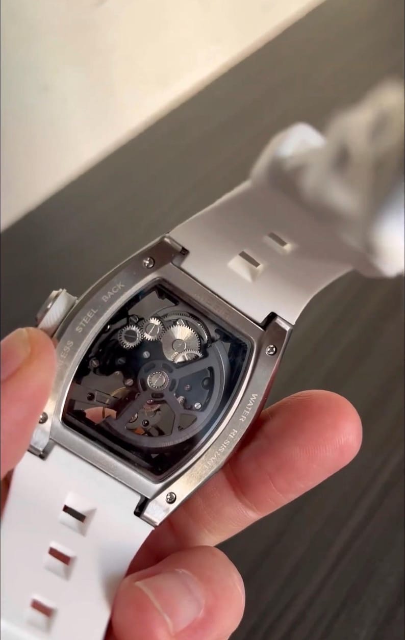 May include: A skeleton watch with a white silicone strap. The watch features a transparent face, revealing the intricate inner workings of the mechanism. The watch case is silver-toned, and the back is marked "Stainless Steel Back" and "Water Resistant".