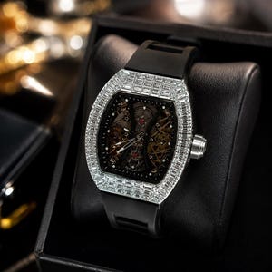 Könnte beinhalten: Eine automatische Armbanduhr mit einem tonnenförmigen, silberfarbenen Gehäuse, das mit klaren Steinen im Baguetteschliff besetzt ist. Die Uhr hat ein schwarzes Skelett-Zifferblatt, das mechanische Komponenten zeigt, und ein glattes schwarzes Kautschukarmband.