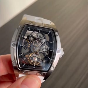 May include: A silver-toned skeleton watch with a white strap. The watch face is black with visible gears and the brand name "MEGIR". The watch has a rectangular case with rounded edges and a white strap.