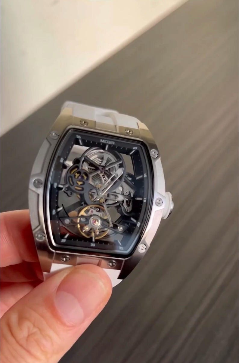 May include: A silver-toned skeleton watch with a white strap. The watch face is black with visible gears and the brand name "MEGIR". The watch has a rectangular case with rounded edges and a white strap.