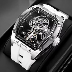 May include: A silver rectangular watch with a black face and a white silicone strap. The watch face has a skeleton design, revealing the inner workings. The watch is displayed on a black surface.
