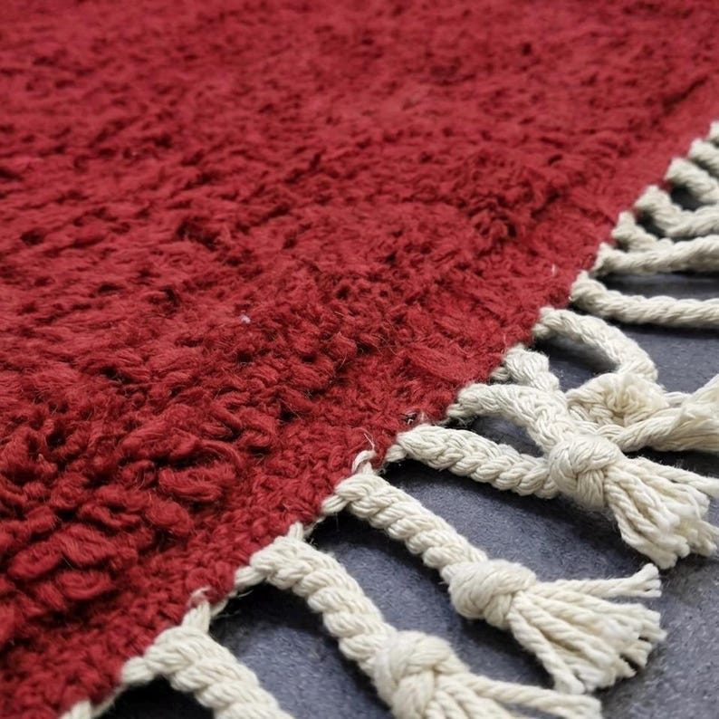 Pu&ograve; includere: Primo piano di un tappeto rosso strutturato con frange color crema. Il tappeto ha un aspetto morbido e ispido. Le frange sono realizzate con spessi cordoncini ritorti, che aggiungono un elemento decorativo al bordo del tappeto. Il tappeto &egrave; probabilmente realizzato in un materiale morbido.