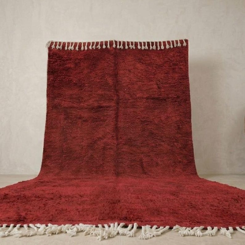 Pu&ograve; includere: Un grande tappeto rettangolare di colore rosso intenso, con una trama a pelo lungo. Il tappeto presenta una frangia di nappe bianche lungo i bordi. Il tappeto &egrave; esposto su una superficie di colore neutro.