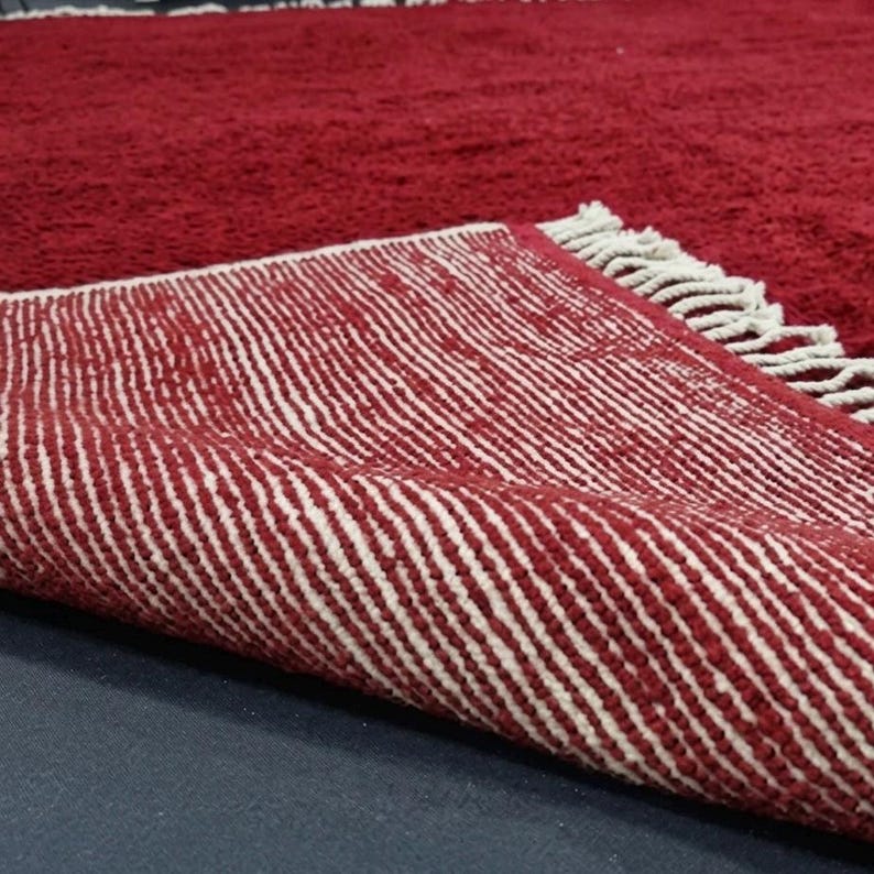Pu&ograve; includere: Primo piano di un tappeto rosso con un motivo a righe bianche sul lato inferiore. Il tappeto ha una consistenza spessa e morbida e una frangia lungo il bordo. Il tappeto &egrave; ripiegato, rivelando il motivo a contrasto.