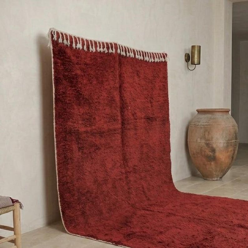 Pu&ograve; includere: Un grande tappeto rosso rettangolare con una trama a pelo lungo. Il tappeto ha una frangia bianca lungo il bordo superiore ed &egrave; drappeggiato contro un muro di colore chiaro. Il tappeto &egrave; parzialmente sul pavimento, creando un effetto fluido. Un vaso decorativo &egrave; sullo sfondo.