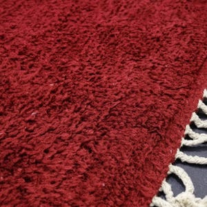 Pu&ograve; includere: Primo piano di un tappeto bordeaux soffice con una superficie strutturata. Il tappeto ha un pelo spesso e una frangia di nappe color crema lungo un bordo. Il colore rosso intenso del tappeto aggiunge calore e consistenza.