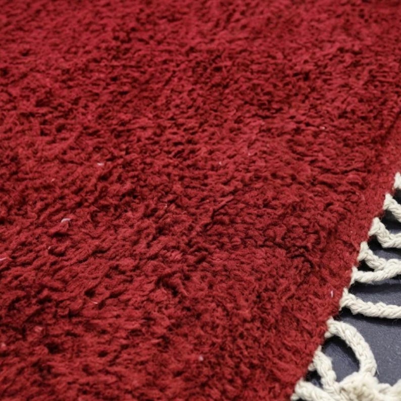 Pu&ograve; includere: Primo piano di un tappeto bordeaux soffice con una superficie strutturata. Il tappeto ha un pelo spesso e una frangia di nappe color crema lungo un bordo. Il colore rosso intenso del tappeto aggiunge calore e consistenza.