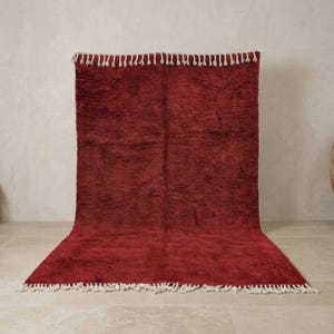 Pu&ograve; includere: Un grande tappeto rettangolare color bordeaux con un fitto pelo strutturato. Il tappeto ha una frangia di nappe bianche lungo i bordi superiore e inferiore. Il tappeto &egrave; esposto contro una parete e un pavimento dai toni neutri.