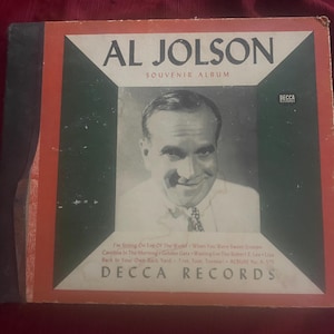 May include: A vintage Al Jolson Souvenir Album by Decca Records. The square cover features a black and white portrait of Al Jolson framed in dark green and orange. Text includes song titles, the Decca Records logo, and album number A-575.