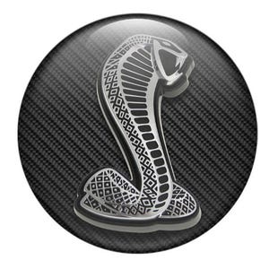 May include: A glossy, circular emblem featuring a silver cobra design on a carbon fiber background. The cobra is detailed with intricate patterns and is set against a textured, dark gray backdrop, creating a striking contrast.