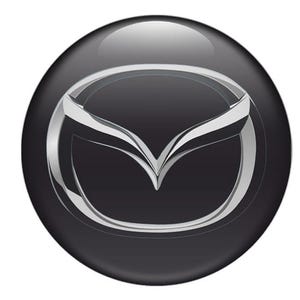 Puede incluir: Un emblema circular negro brillante con el logotipo de Mazda. El logotipo es un emblema plateado estilizado con un diseño en forma de ala, sobre un fondo oscuro. El emblema tiene una apariencia reflectante y tridimensional.
