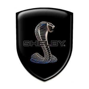 May include: A black shield-shaped emblem featuring a silver cobra with blue accents. The word "SHELBY" is printed in white below the snake. The emblem has a glossy finish and is set against a white background.