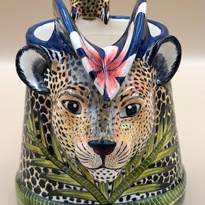 May include: A ceramic pitcher with a leopard face design, featuring a spotted pattern in tan and black. The pitcher has blue accents, a pink and white flower, and a small bird on top. Green leaves and a green base complete the design.