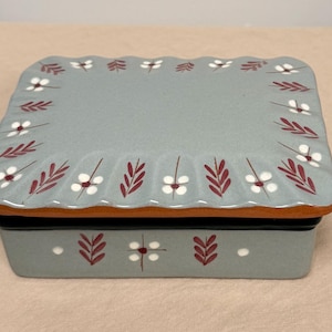 May include: A rectangular ceramic lidded box with a scalloped edge on the lid. The box is light blue-gray with a hand-painted pattern of small white flowers and dark red leaf sprigs. The base of the lid shows a natural terracotta-colored rim.