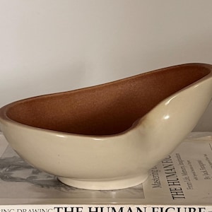 May include: An asymmetrical ceramic bowl with a smooth, glossy off-white exterior and a contrasting matte, terracotta-colored interior. The bowl features a curved, elongated rim that tapers to a point on one side, resting on a circular base.