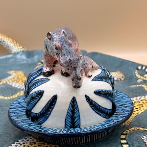 May include: A ceramic butter dish featuring a hippo and calf figurine on the lid. The lid is white with blue leaf-like designs and a dark blue rim. The dish sits on a patterned surface with leopard print.