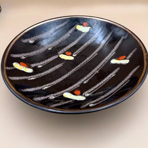 May include: A black ceramic serving bowl with a gold rim. The bowl features a pattern of white diagonal lines and small yellow and red abstract designs. The bowl is round and has a slightly raised edge.