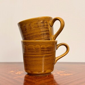 May include: Two stacked ceramic mugs in a warm ochre brown tone, featuring a horizontal brushed texture and a scalloped rim detail. Each mug has a curved handle on the side, with the top mug resting directly on the rim of the bottom mug.