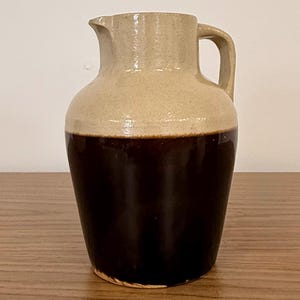 May include: A two-toned ceramic pitcher featuring a glossy dark brown glaze on the lower half and a matte light beige finish on the upper section. The vessel has a rounded body, a wide neck, a small pouring spout, and a sturdy side handle.