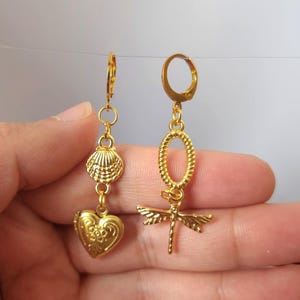 May include: Gold-colored dangle earrings. One earring features a seashell and heart charm, the other an oval and dragonfly charm. The earrings have a lever-back closure and are made of a shiny, metallic material.