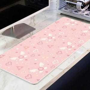 May include: A rectangular pink desk mat featuring a repeating pattern of winged circles, hearts, triangles, and diamonds. The mat has a white border and is placed on a marble-patterned surface.