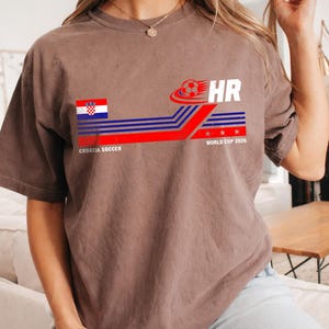 May include: A brown short-sleeve t-shirt featuring a graphic with the Croatian flag, a soccer ball, the text HR, and the words CROATIA SOCCER WORLD CUP 2026. The design includes horizontal blue and red stripes with three small white stars.