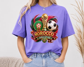 May include: A light purple t-shirt featuring a graphic print of the Morocco World Cup 2026 logo, a gold trophy, a soccer ball, and the Moroccan national emblem, all set against a swirling red and green background with the text Morocco World Cup 2026.