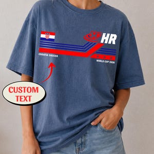 May include: A blue short-sleeve t-shirt featuring a graphic with the Croatian flag, the text "CROATIA SOCCER", "HR", and "WORLD CUP 2026" alongside a soccer ball icon. A red arrow points to the text area with a label reading "CUSTOM TEXT".
