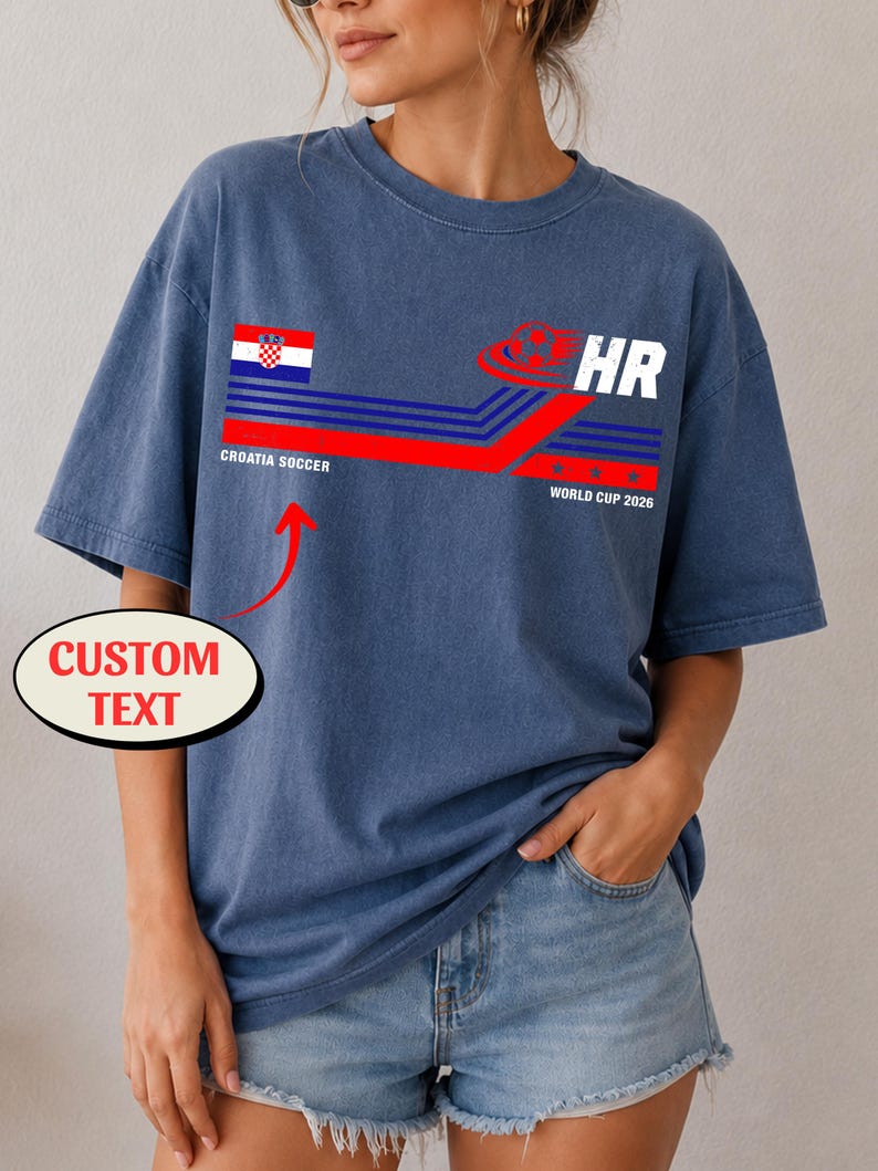 May include: A blue short-sleeve t-shirt featuring a graphic with the Croatian flag, the text "CROATIA SOCCER", "HR", and "WORLD CUP 2026" alongside a soccer ball icon. A red arrow points to the text area with a label reading "CUSTOM TEXT".