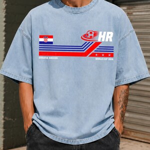 May include: A light blue short-sleeve t-shirt featuring a graphic with the Croatian flag, a soccer ball, the letters HR, and the text CROATIA SOCCER WORLD CUP 2026. The design includes horizontal blue and red stripes with three white stars.