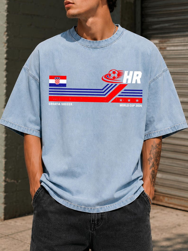 May include: A light blue short-sleeve t-shirt featuring a graphic with the Croatian flag, a soccer ball, the letters HR, and the text CROATIA SOCCER WORLD CUP 2026. The design includes horizontal blue and red stripes with three white stars.