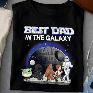以下が含まれることがあります： 白と青で「BEST DAD IN THE GALAXY」の文字が入った黒いTシャツ。ダース・ベイダー、チューバッカ、ストームトルーパーなど、スター・ウォーズのキャラクターのグラフィックが特徴です。キャラクターの下に名前がプリントされています。