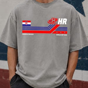 May include: A grey short-sleeve t-shirt featuring a graphic with the Croatian flag, a soccer ball, and the text "HR", "CROATIA SOCCER", and "WORLD CUP 2026". The design includes horizontal blue and red stripes with three small stars.