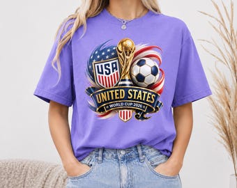 May include: A light purple t-shirt featuring a graphic of the United States World Cup 2026 logo, a gold trophy, a soccer ball, and a shield with the text USA, United States, and World Cup 2026, all surrounded by red, white, and blue swirls.