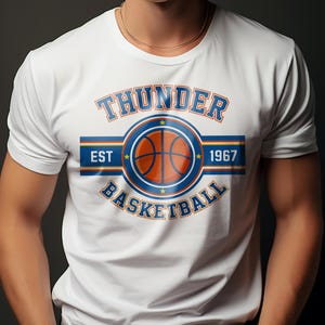 Op de afbeelding: Wit T-shirt met een basketbalontwerp. Het ontwerp bevat het woord "THUNDER" in blauw, het woord "BASKETBALL" in blauw en een oranje en blauwe basketbalafbeelding. De tekst "EST 1967" is ook opgenomen.